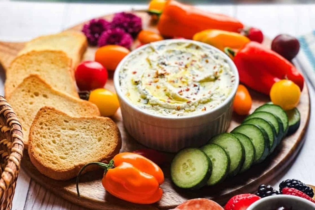 A vibrant platter with fresh vegetables, sliced bread, and a bowl of dip, attractively arranged on a wooden board as bites for dinner.