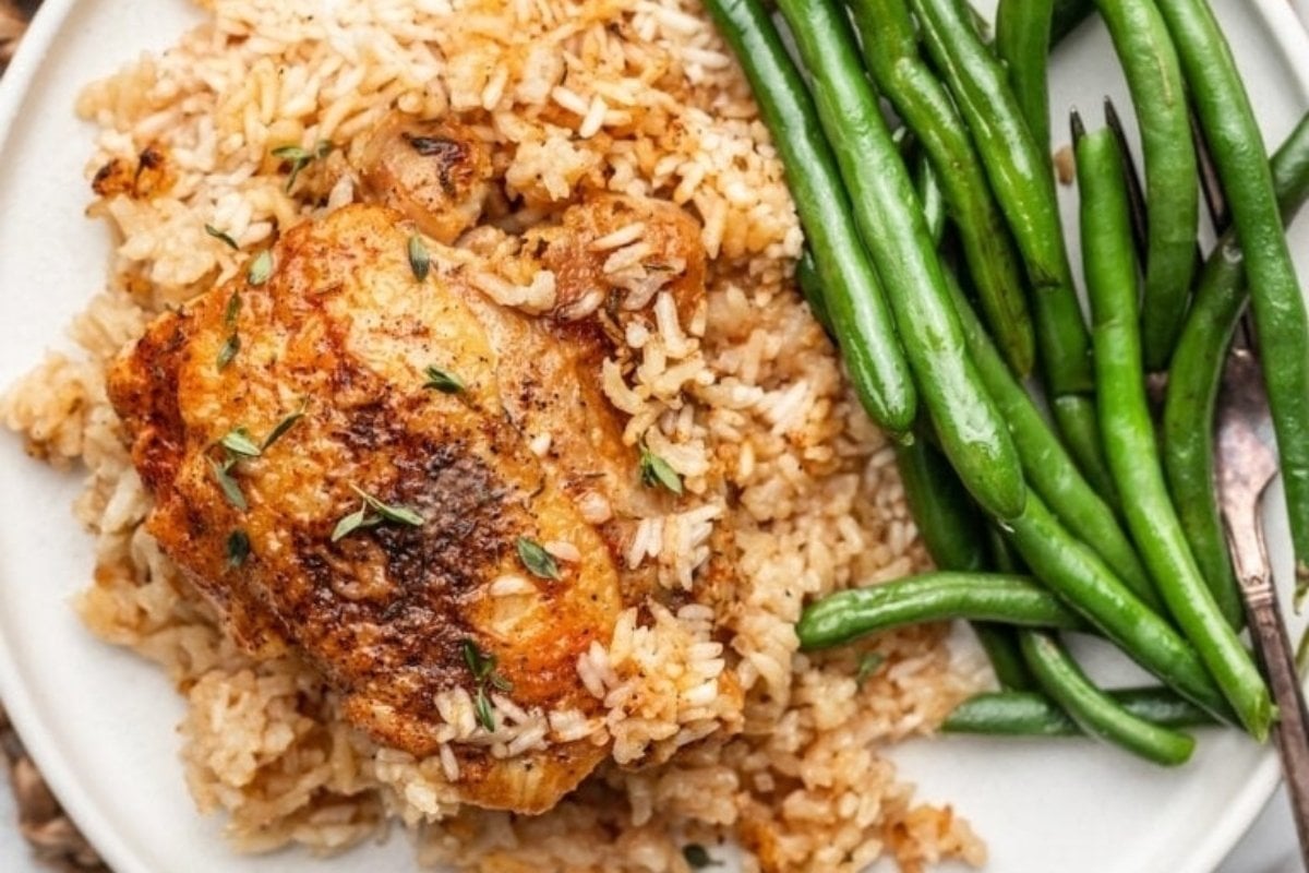 A plate of seasoned chicken thigh served with rice and green beans, garnished with herbs, perfect for comfort food recipes.