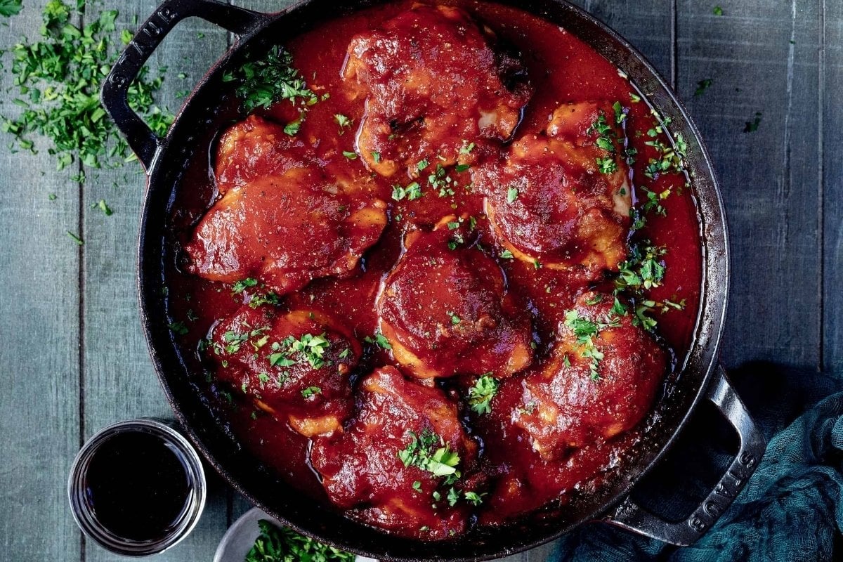 Chicken thighs in rich red tomato sauce, garnished with chopped herbs, simmer in a black skillet on a gray table—ideal for those seeking hearty comfort food recipes.