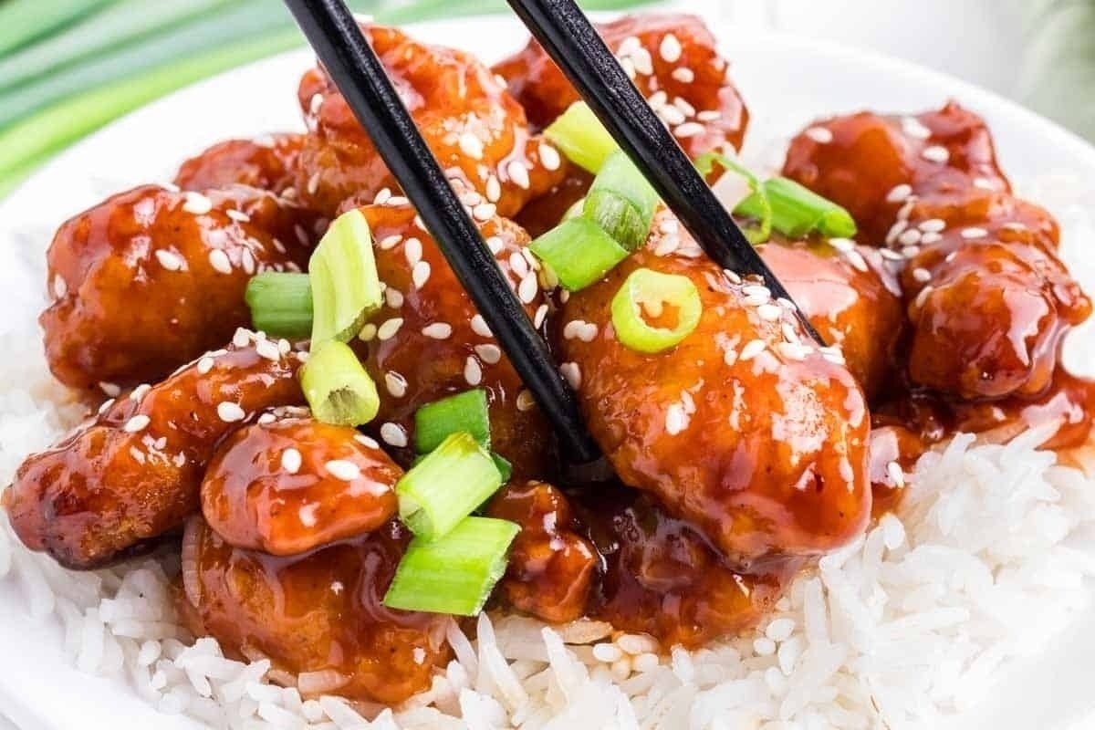 Chopsticks lifting a piece of sesame chicken coated in a sticky sauce, served over white rice with chopped green onions. Ideal for comfort food recipes.
