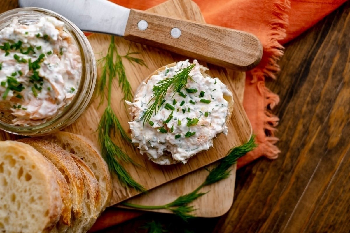 A jar of salmon spread with chives, a spread-covered slice of bread, and a knife on a wooden board next to sliced bread and an orange napkin, perfect for bites for dinner.