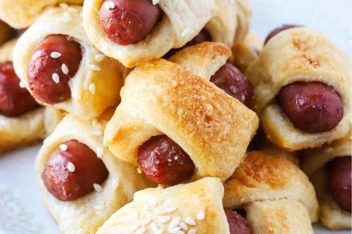 Pigs in a blanket; small sausages wrapped in golden-brown, flaky pastry, sprinkled with sesame seeds on a white plate, perfect as bites for dinner.