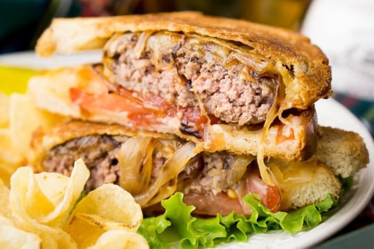 A classic comfort food, this patty melt sandwich comes with grilled onions, tomato, and melted cheese, served alongside crisp lettuce and potato chips.