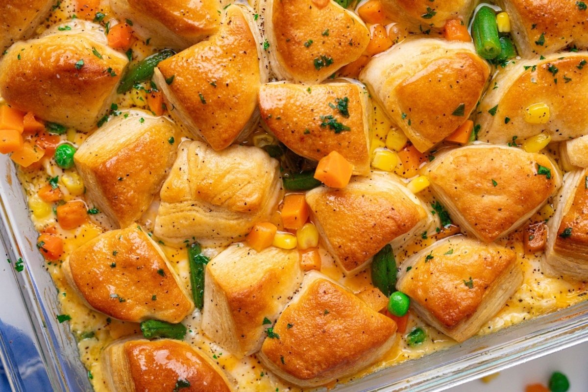 A glass baking dish filled with baked dinner rolls surrounded by mixed vegetables in a creamy sauce, garnished with parsley, epitomizing comfort food.