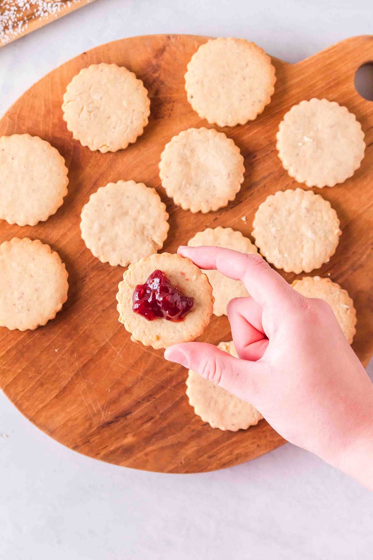 A hand holds a Linzer cookie filled with red jam; more plain cookies are arranged on a round wooden board.