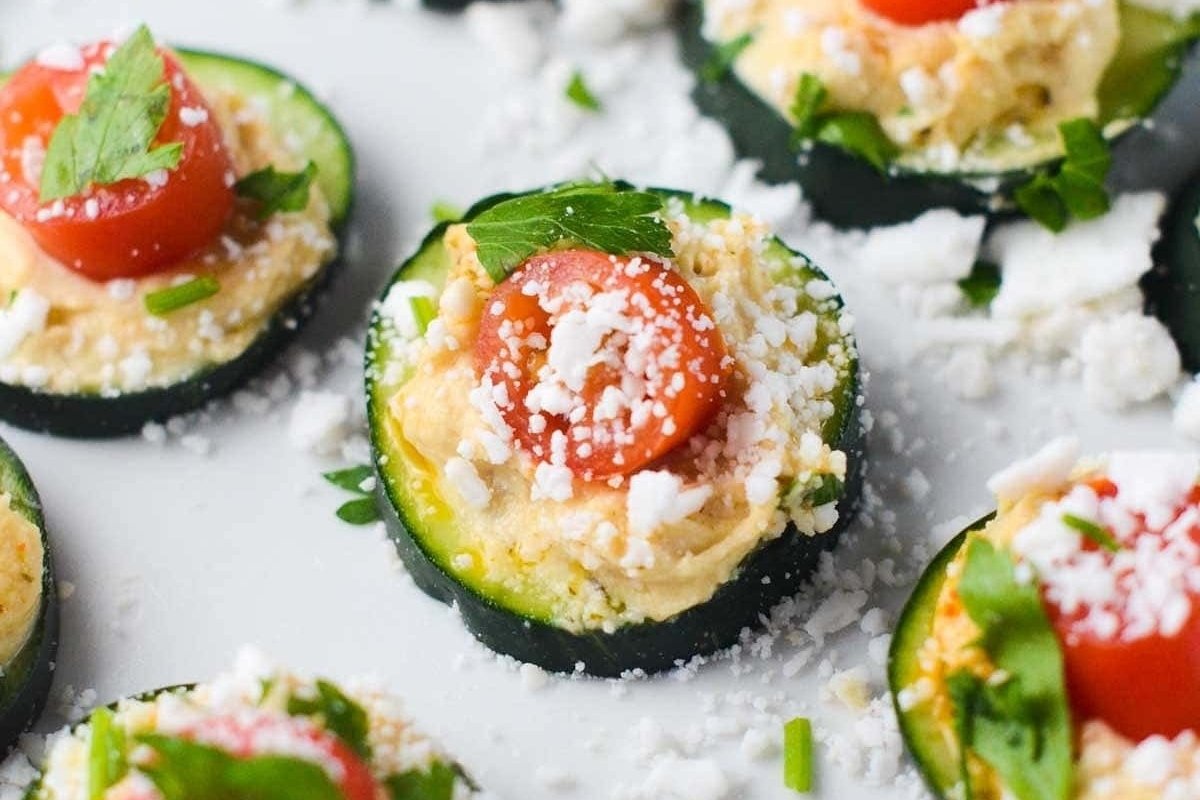 Slices of zucchini topped with hummus, cherry tomatoes, feta cheese, and fresh basil on a white surface, perfect as bites for dinner.