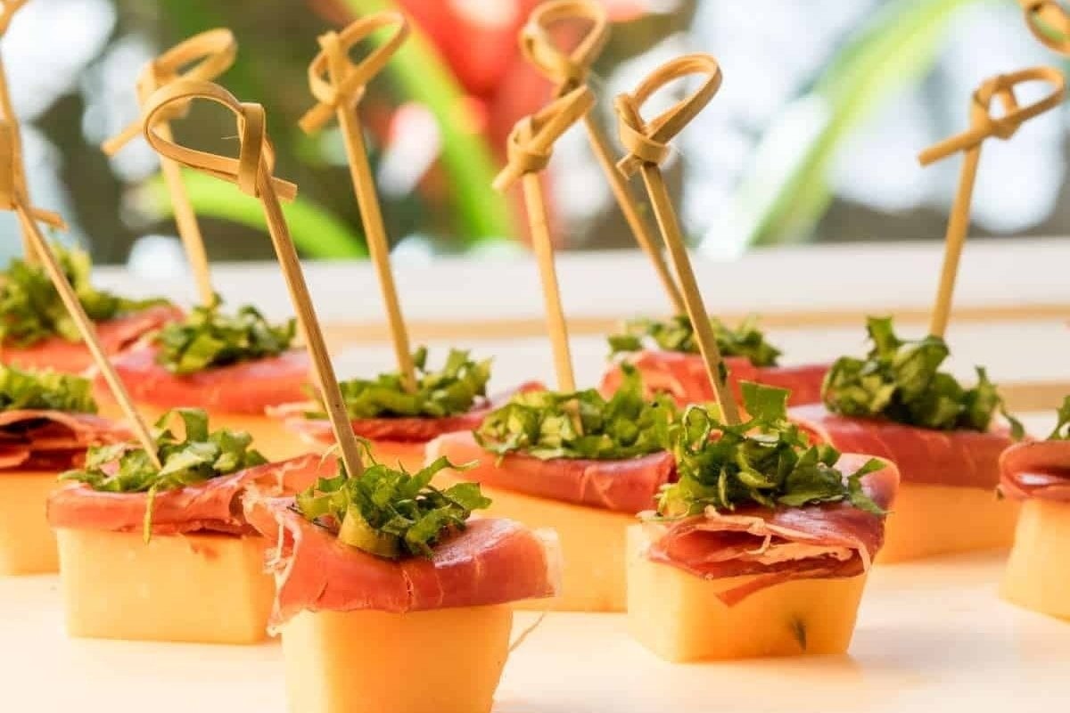 Appetizers featuring prosciutto and garnished herbs on small bread slices, secured with decorative toothpicks, presented as bites for dinner on a tray.