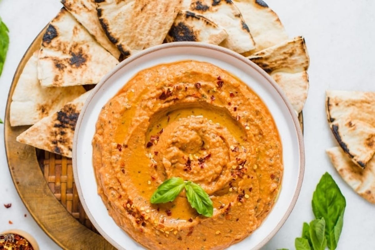 A bowl of creamy roasted red pepper hummus topped with basil, surrounded by bites for dinner of grilled pita bread on a plate.