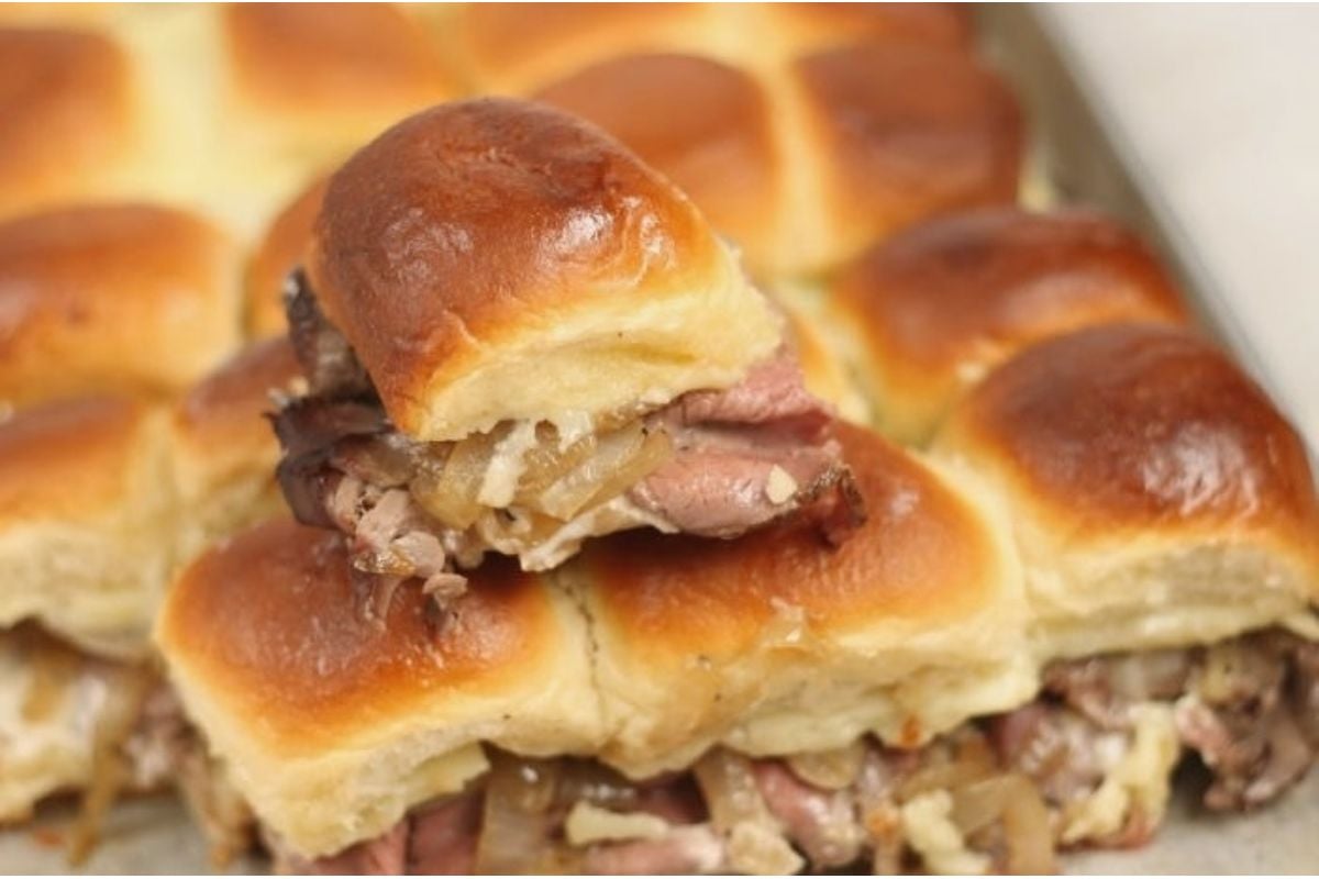 Close-up of game day sliders filled with sliced meat and caramelized onions, stacked bite size on a tray.