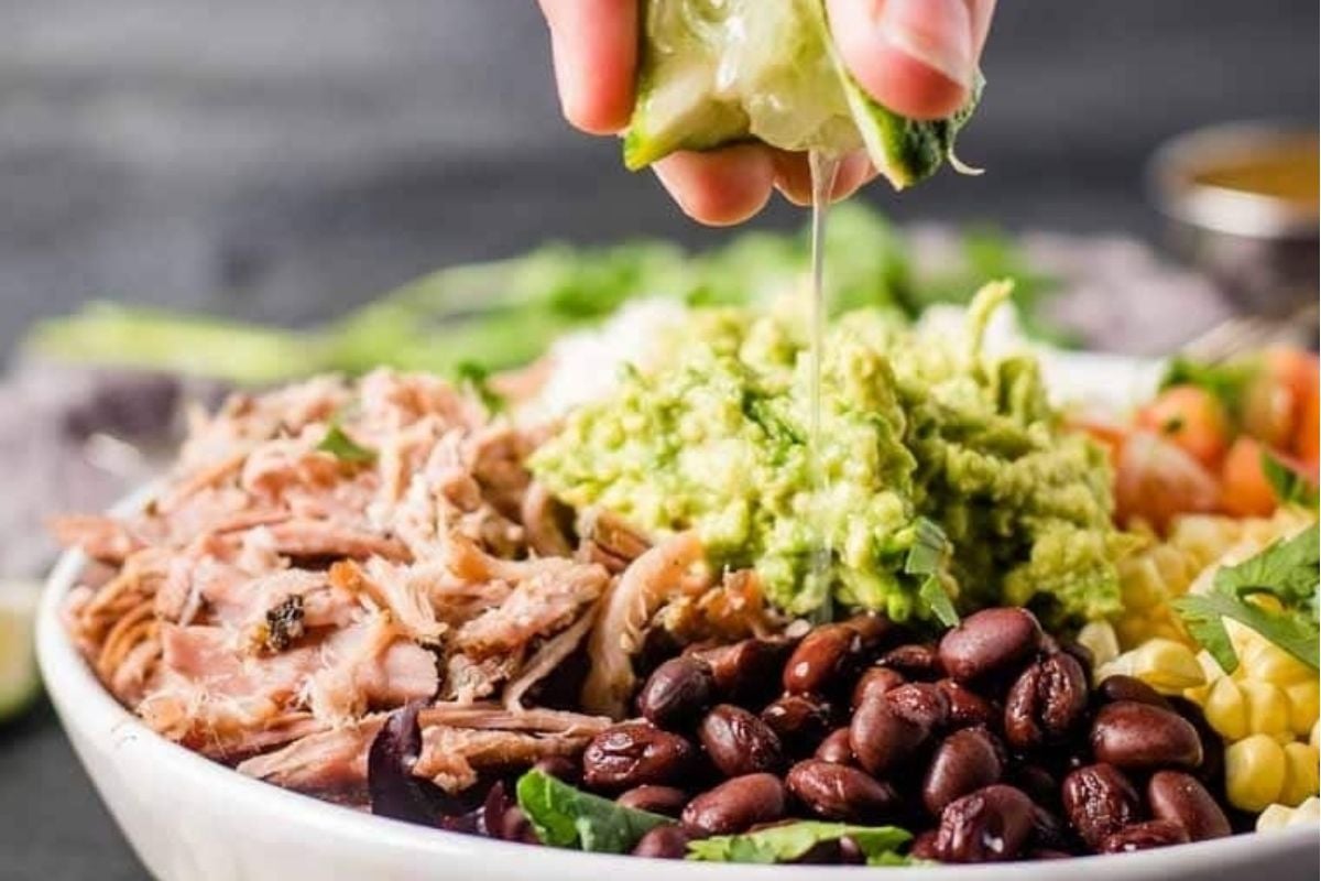 A hand squeezes lime over a bowl with slow cooker game day recipes like pulled pork, black beans, guacamole, corn, and vegetables.