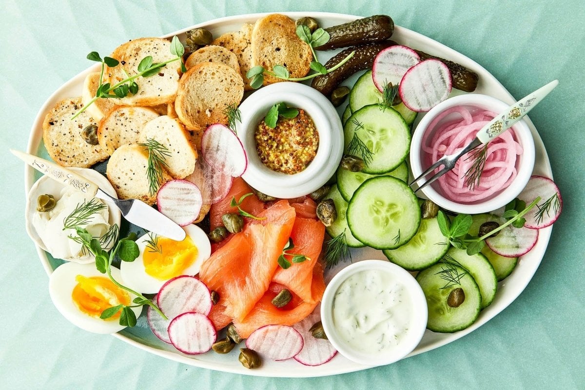 A platter of smoked salmon, assorted vegetables, bread, pickles, and condiments, garnished with herbs and perfect as bites for dinner.