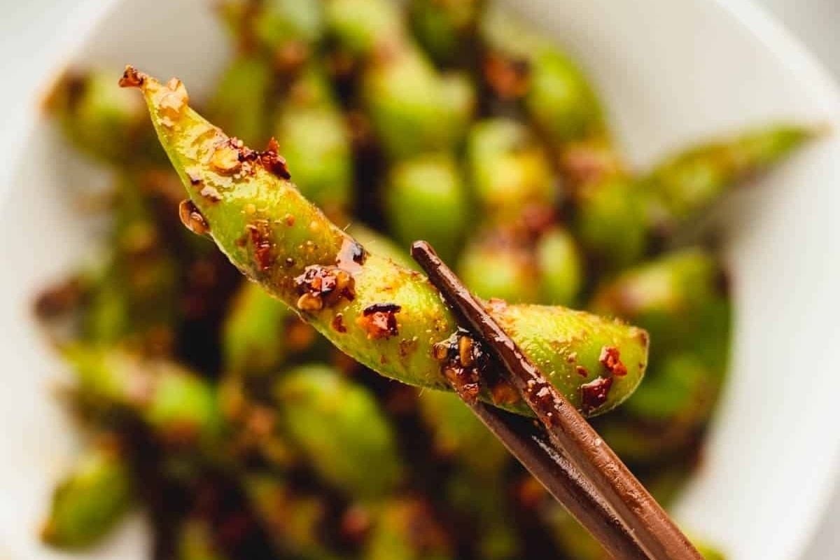 A pair of chopsticks holds a bite-size seasoned green bean over a white bowl filled with more cooked green beans topped with red chili flakes.