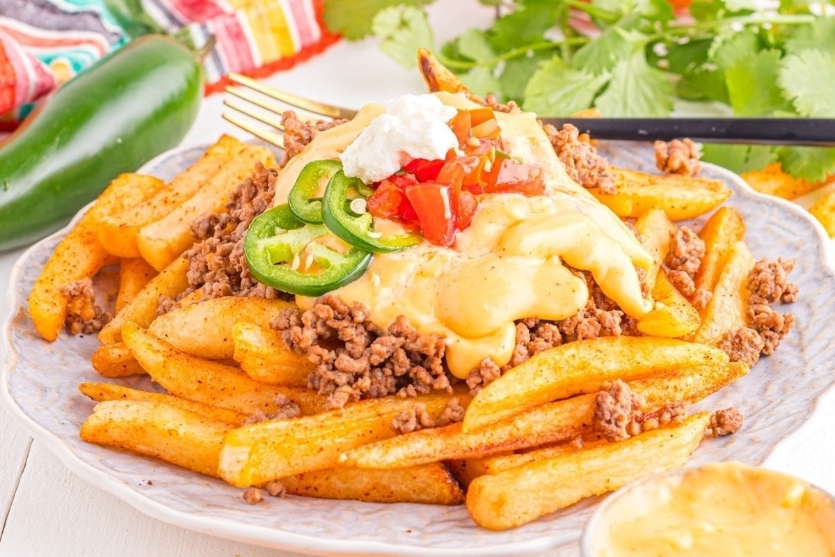 A plate of seasoned fries topped with ground beef, melted cheese, jalapeños, dollops of sour cream, and diced tomatoes, served with a side of cheese sauce— a classic from comfort food