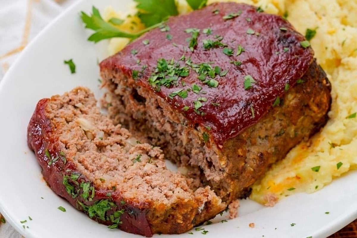 A slice of meatloaf, a classic from comfort food recipes, topped with a glossy sauce, garnished with chopped parsley, served on a white plate with mashed potatoes.