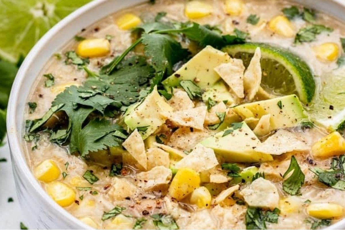Creamy soup with corn, avocado slices, tortilla chips, cilantro, and lime in a white bowl—perfect for slow cooker game day recipes.