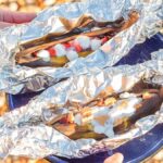 A person is holding two foil-wrapped campfire banana boats on a plate.
