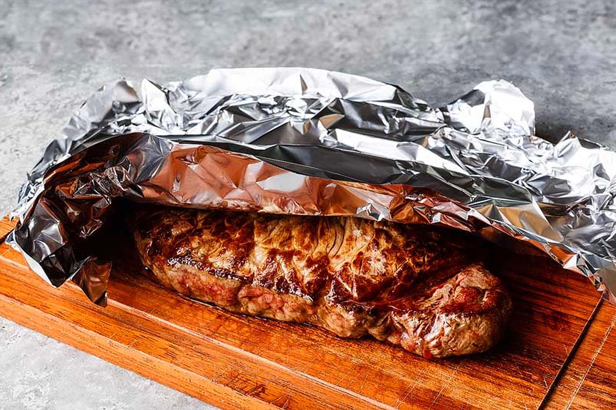 Grilled steak resting on wooden board covered with aluminum foil