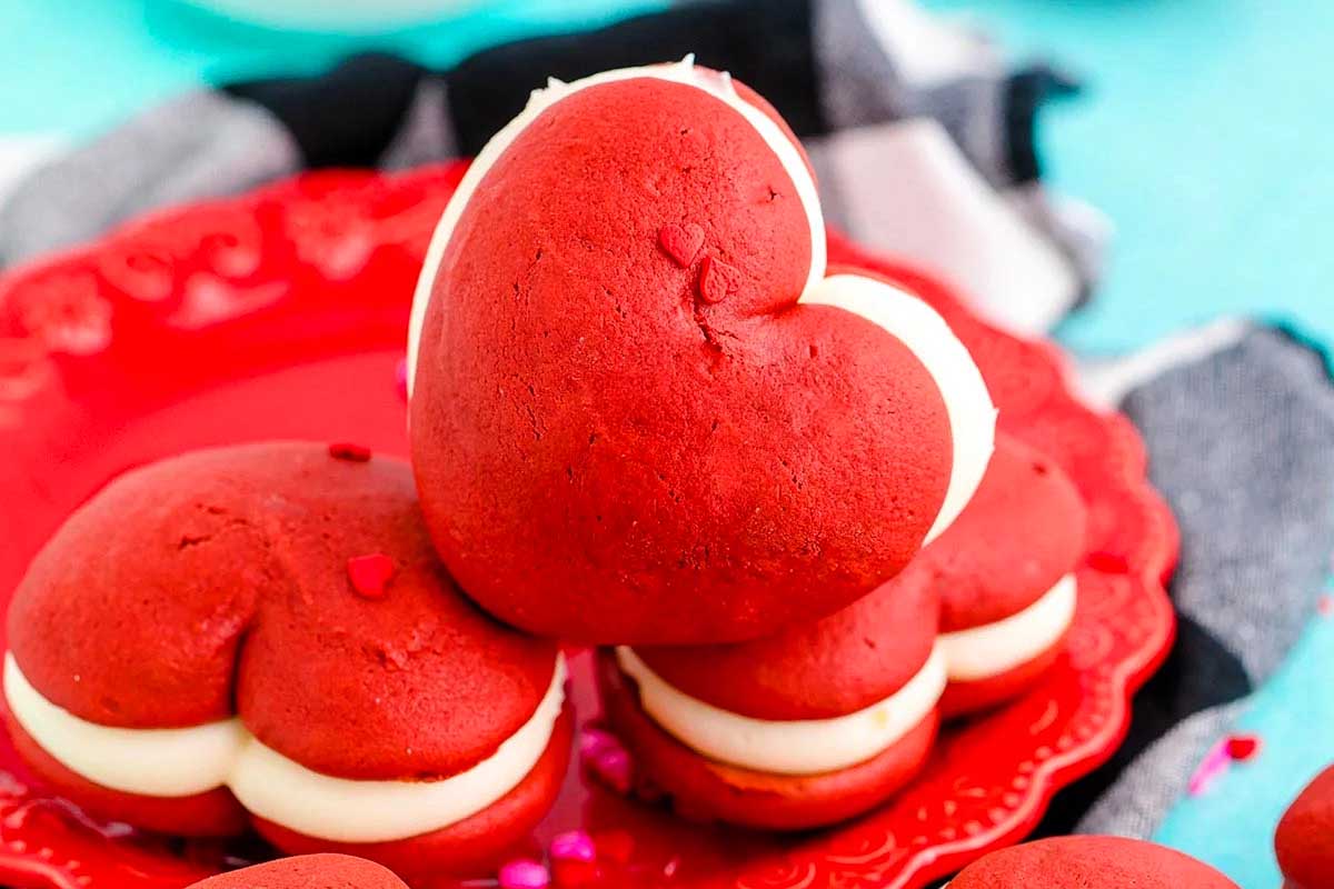 Heart-shaped red velvet whoopie pies with white cream filling on a red plate—perfect Valentine's Day cookies for sharing with someone special.