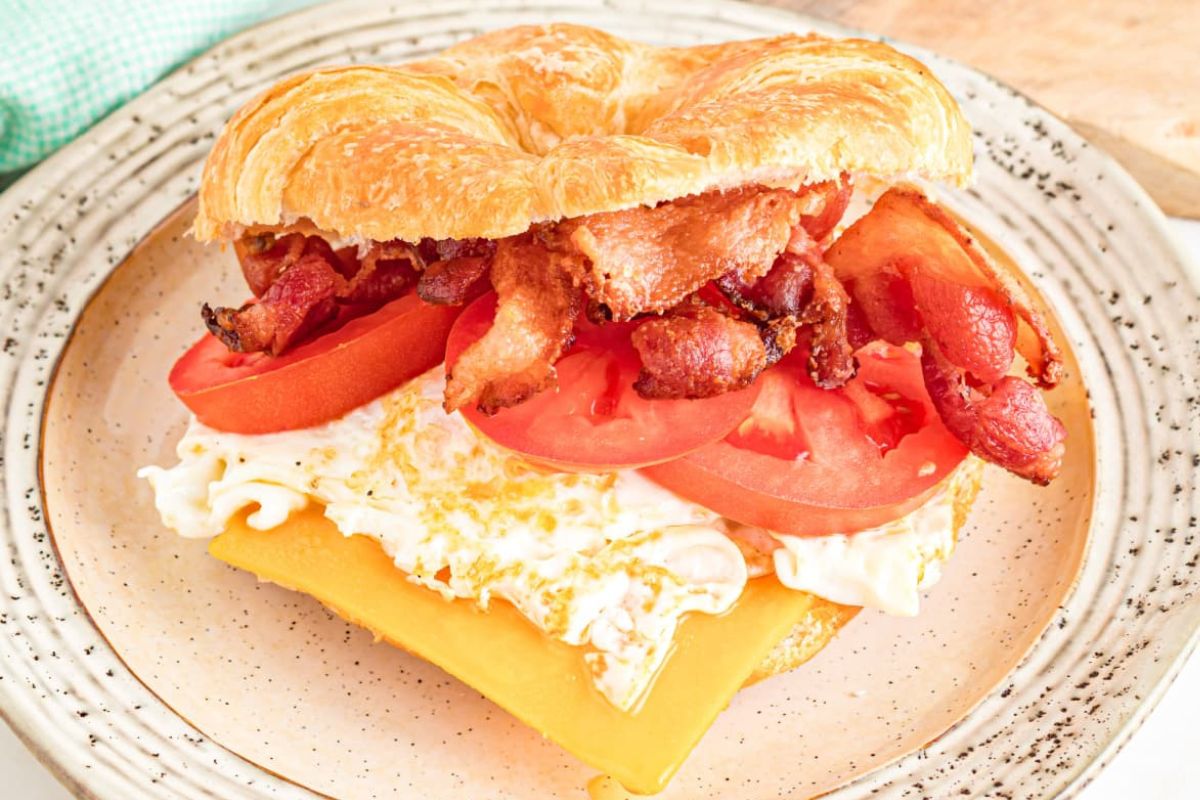 A croissant sandwich with bacon, tomatoes, cheese, and egg on a plate.