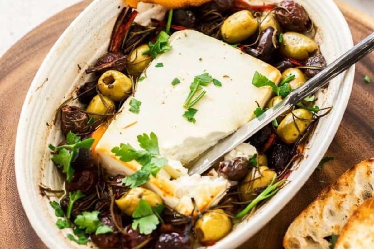 A dish of baked feta cheese from our cheesy recipes, garnished with olives, herbs, and sun-dried tomatoes, served with crusty bread on the side.