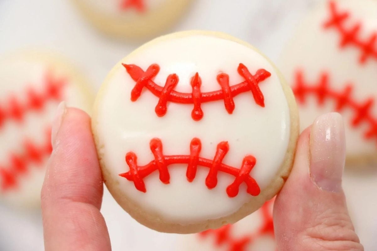 A hand holding a baseball-themed cookie, crafted using one of our unique cookie recipes, with white icing and red stitching details.