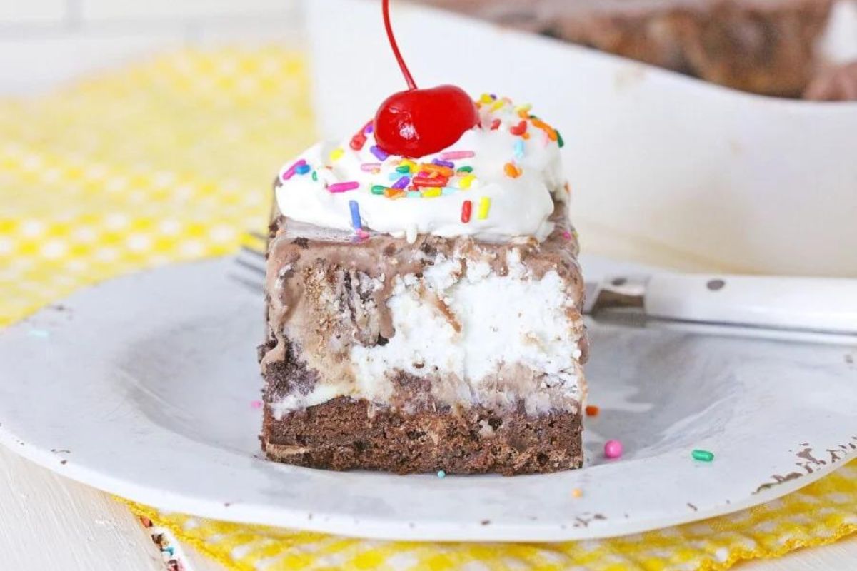 A slice of layered ice cream cake with ice cream recipes, sprinkles, and a cherry on top, displayed on a white plate.