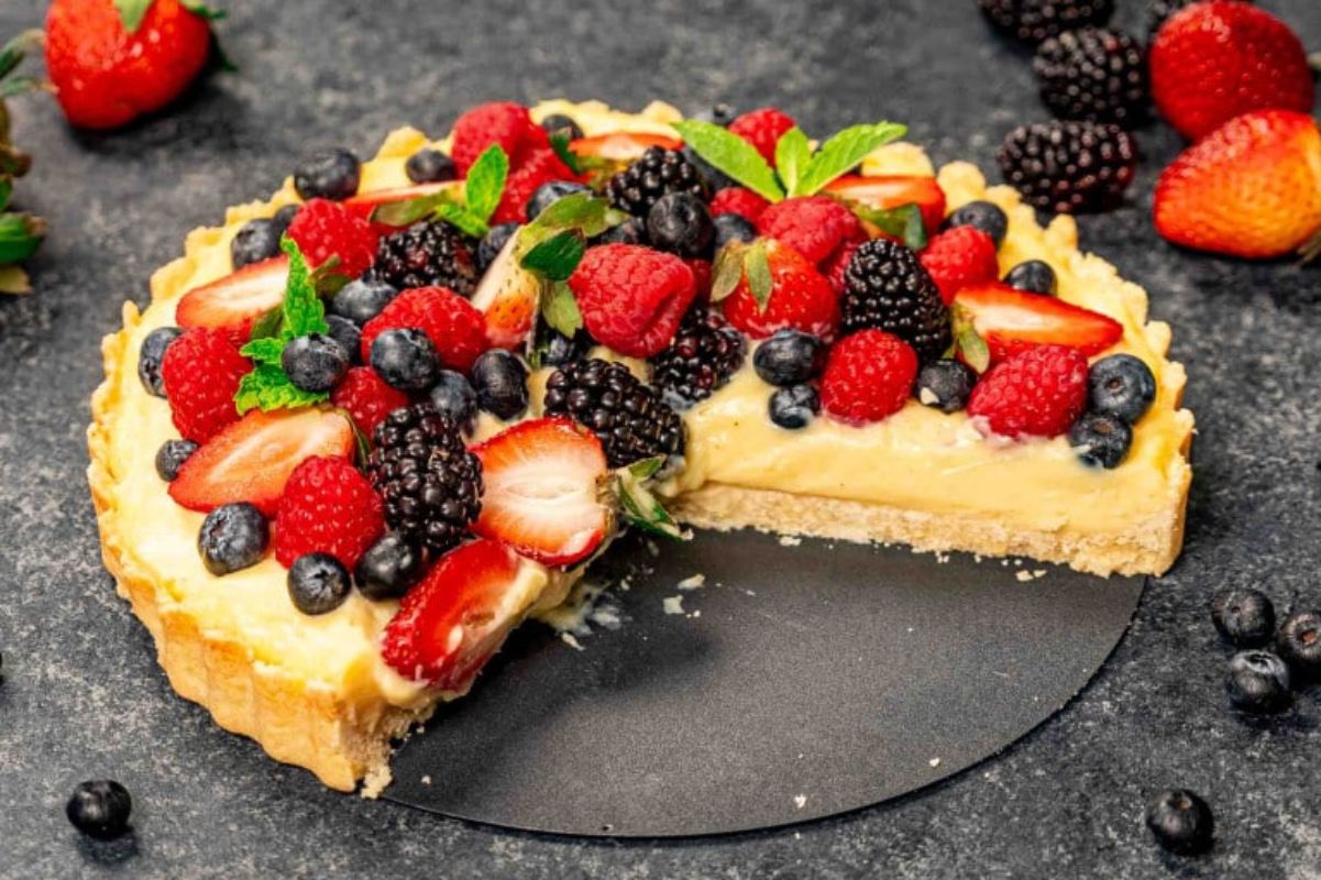 An Easter fresh fruit tart with a slice removed, showcasing an assortment of berries and a creamy filling on a dark surface.
