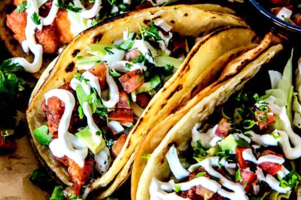 Three tacos with crispy fish, fresh cilantro, diced tomatoes, and drizzled with creamy sauce from our seafood recipes, served on a rustic plate.