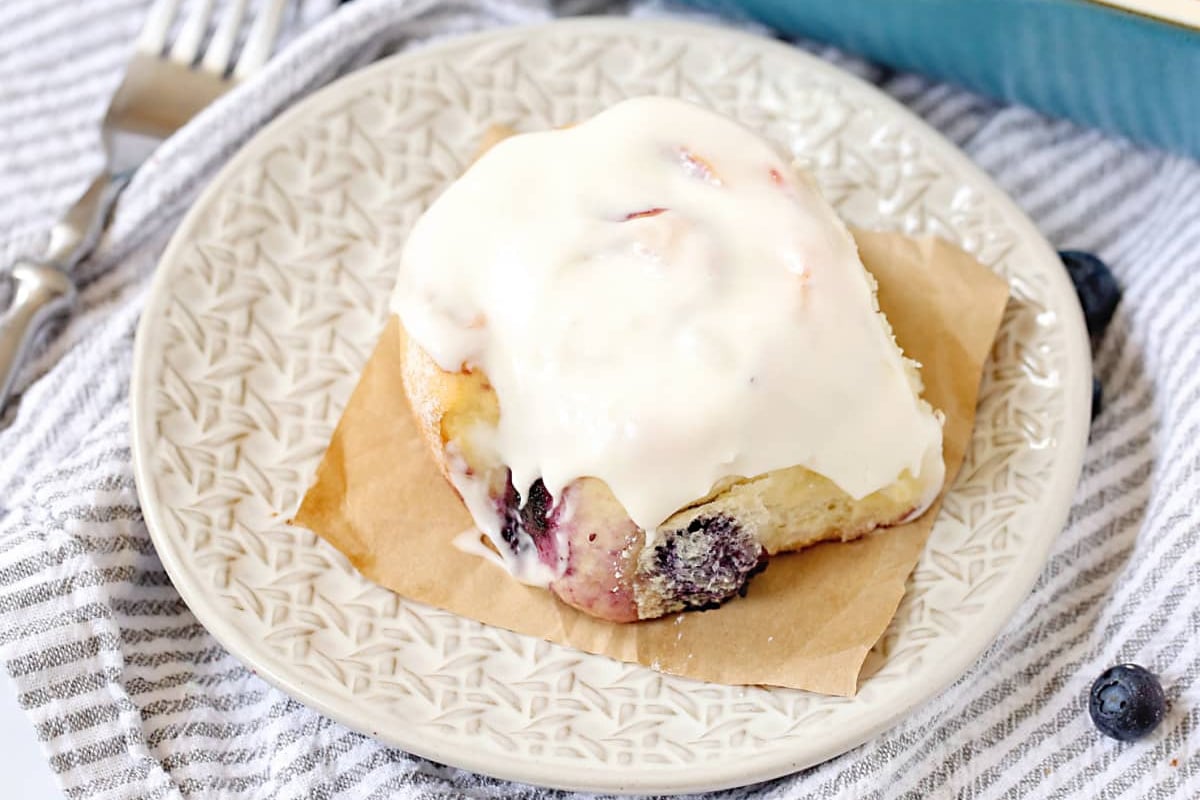 A freshly baked cinnamon roll recipe with white icing and blueberries on a decorative plate.