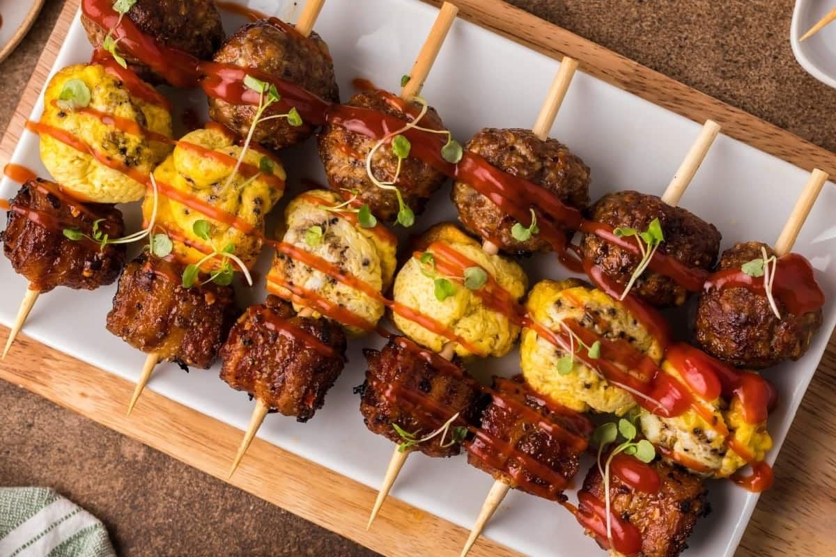 A wooden platter holds skewers of grilled meat, egg, and vegetables drizzled with sauce, garnished with microgreens—perfect for a special Father's Day breakfast.