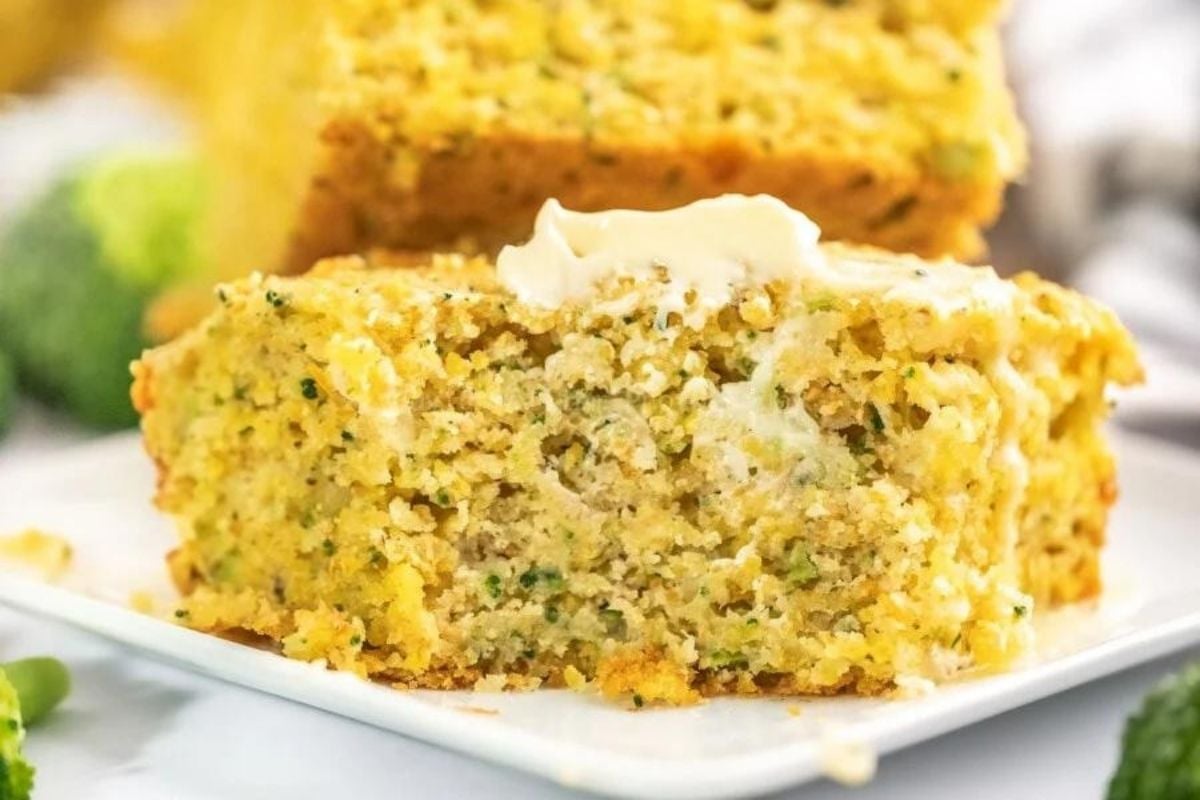 A slice of cheesy broccoli cheddar cornbread topped with a dollop of butter on a white plate.