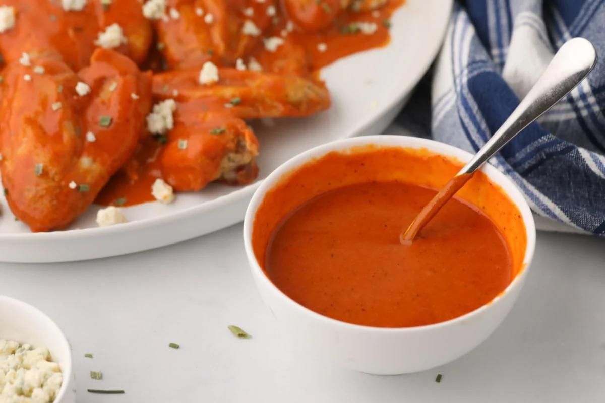 A bowl of buffalo sauce with a spoon, accompanied by a plate of chicken wings and a side of blue cheese crumbles.