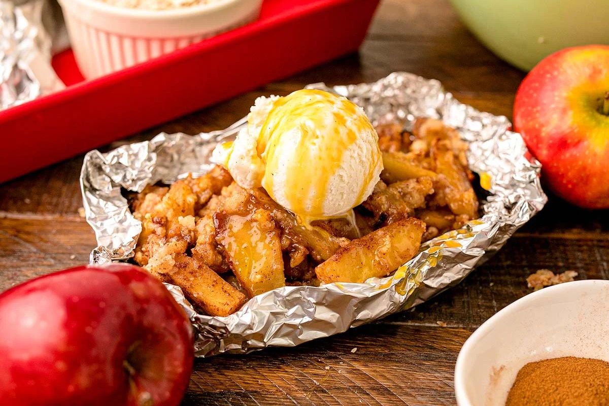 Baked apple crisp in foil, a favorite among campfire recipes, topped with vanilla ice cream and caramel, surrounded by apples and cinnamon.