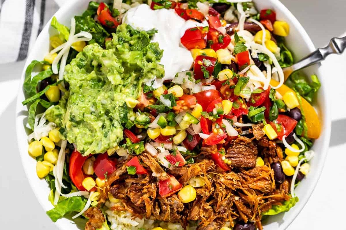 A colorful bowl of salad inspired by burrito recipes, topped with shredded beef, guacamole, sour cream, and fresh vegetables like tomatoes and corn.