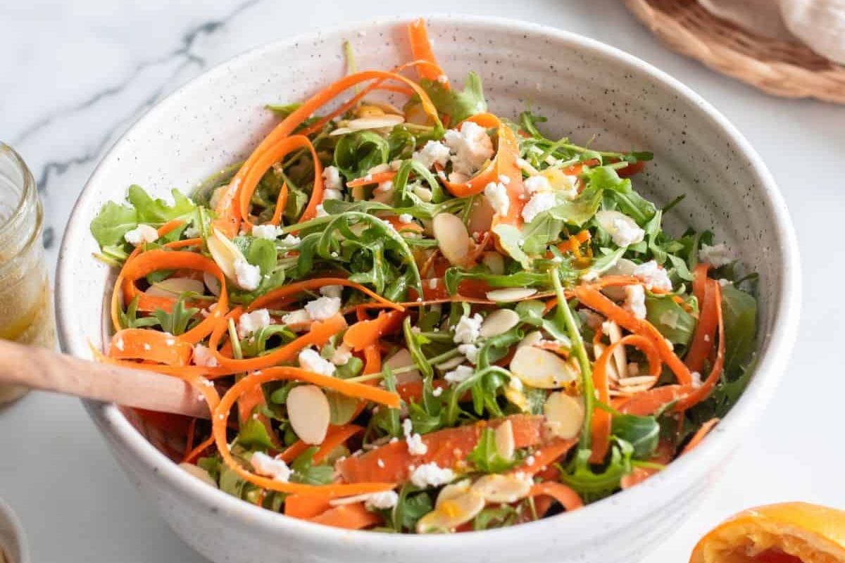 A fresh Easter appetizer salad with arugula, shaved carrots, crumbled cheese, and sliced almonds in a bowl with a wooden spoon.
