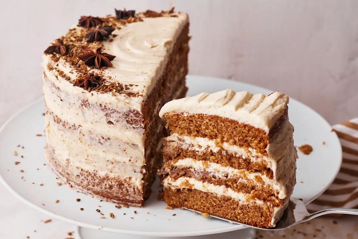 A slice of frosted carrot layer cakes with a piece removed, displaying its layers, on a white plate.