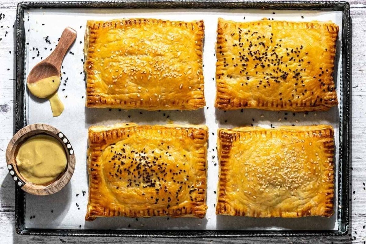 Four cheesy savory pastries with golden-brown crusts topped with sesame and nigella seeds, served on a baking sheet next to small bowls of mustard and sauce.