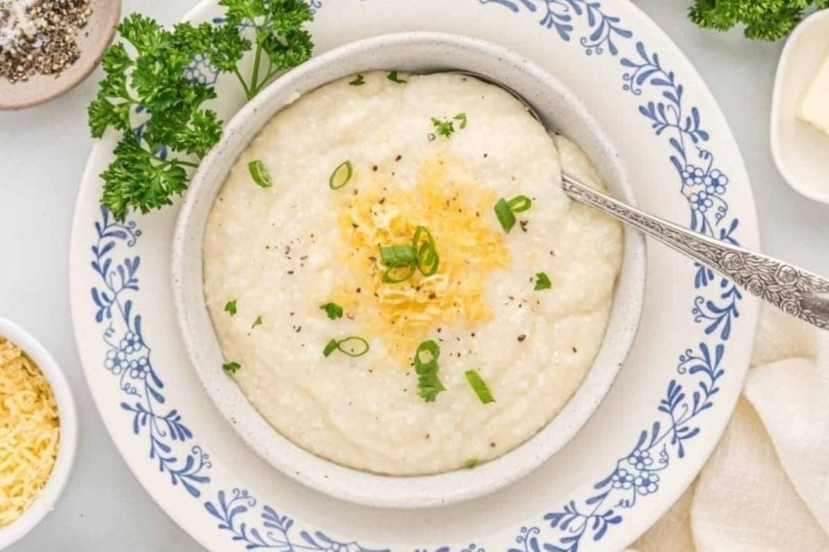 A bowl of creamy grits topped with shredded cheese and green onions, garnished with parsley, on a blue-patterned plate. This is one of our favorite cheesy recipes.