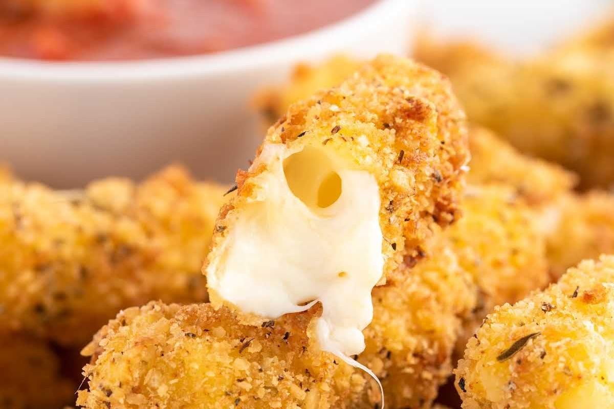 Close-up of a crispy mozzarella stick with gooey melted cheese, perfect for fans of cheesy recipes, with a bowl of marinara sauce in the background.