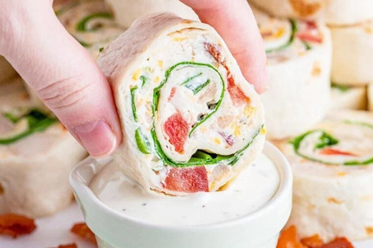 A hand dipping a turkey and vegetable pinwheel into a bowl of sauce.