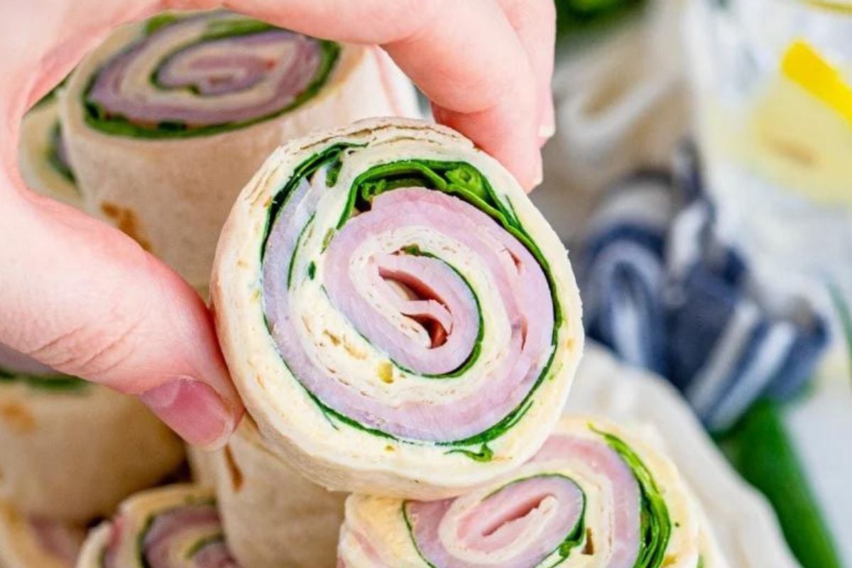 A close-up of a hand holding a pinwheel sandwich from our Pinwheel Recipes collection, featuring turkey, cheese, and spinach wrapped in a tortilla.
