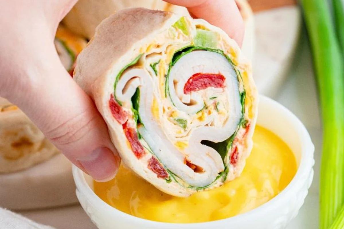 A hand dipping a pinwheel sandwich wrap into a bowl of yellow dip according to pinwheel recipes.