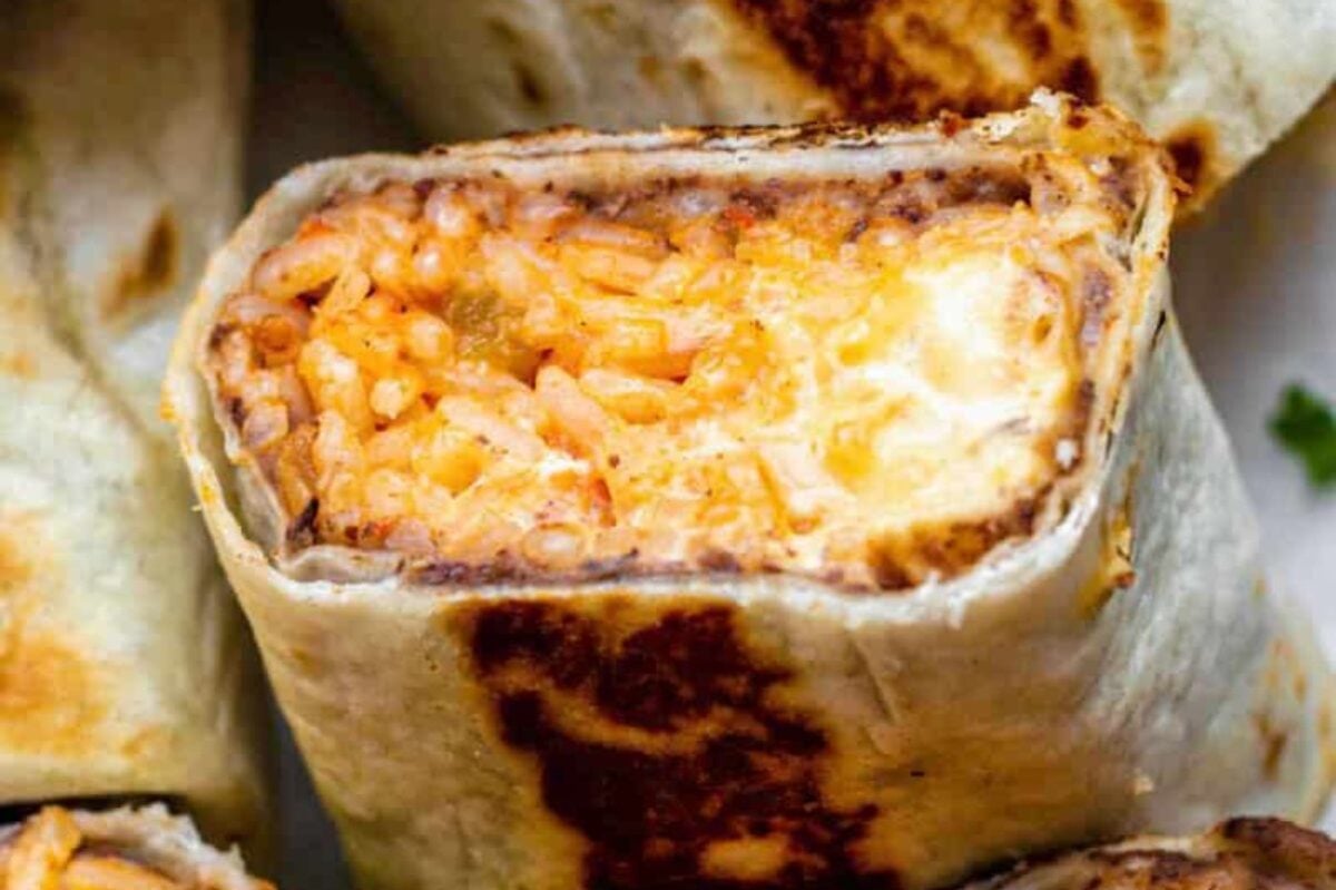 Close-up of a halved burrito from our burrito recipes showing its gooey cheese and rice filling, with another intact burrito and chopped parsley in the background.