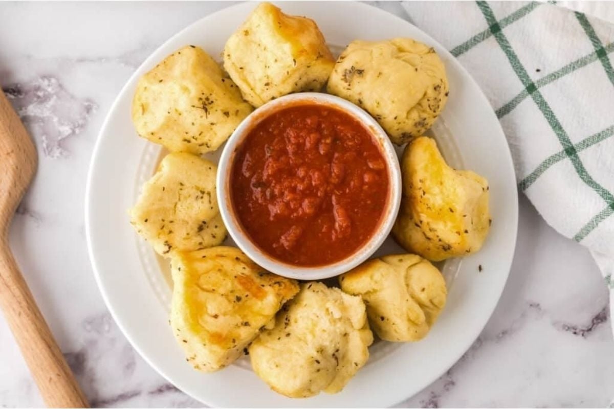 A plate of cheesy garlic knots served with a bowl of marinara sauce, on a marble surface next to a wooden spoon.