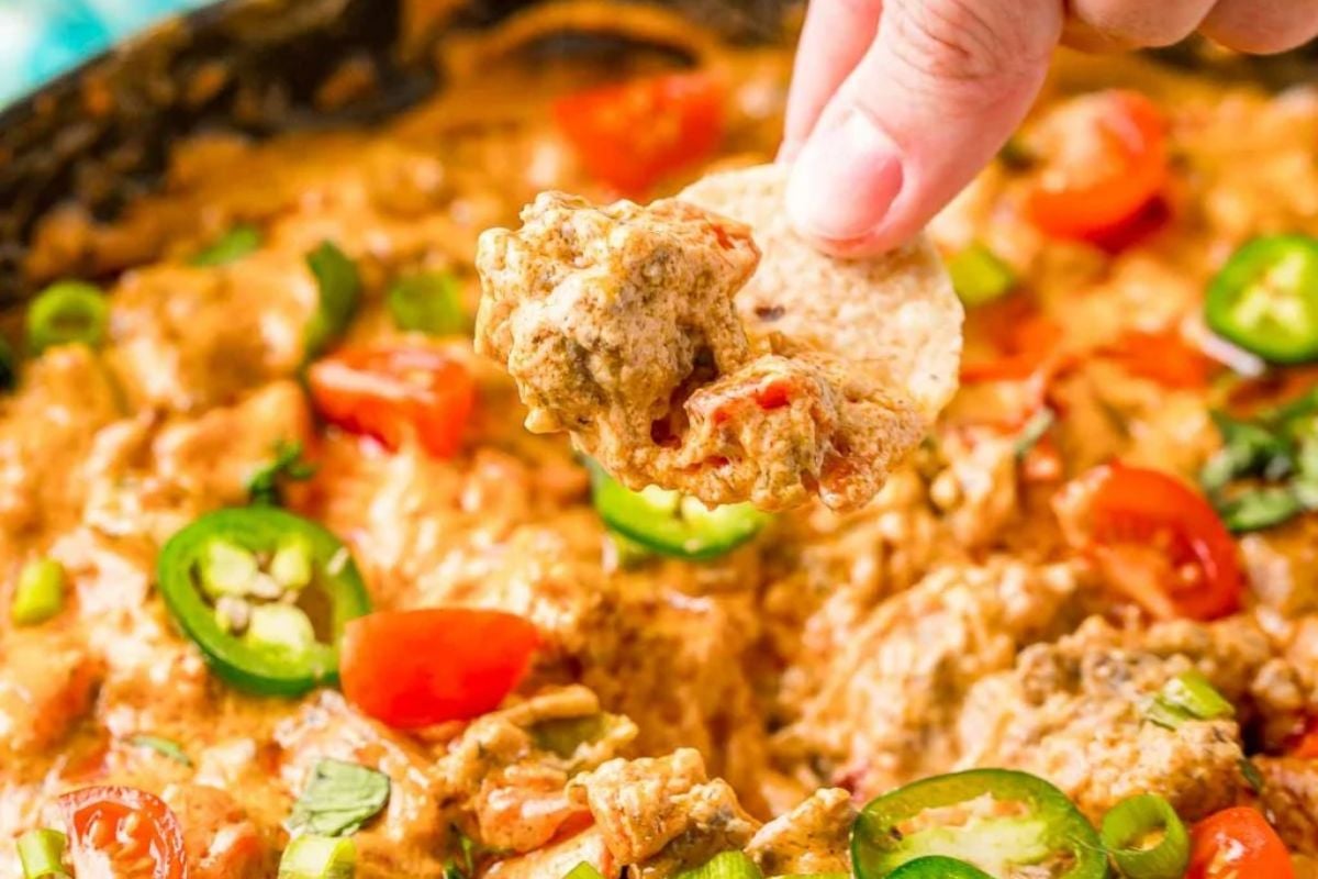 A hand dips a tortilla chip into cheesy, spicy dip topped with tomatoes, jalapeños, and green onions—perfect for fans of cheesy recipes.