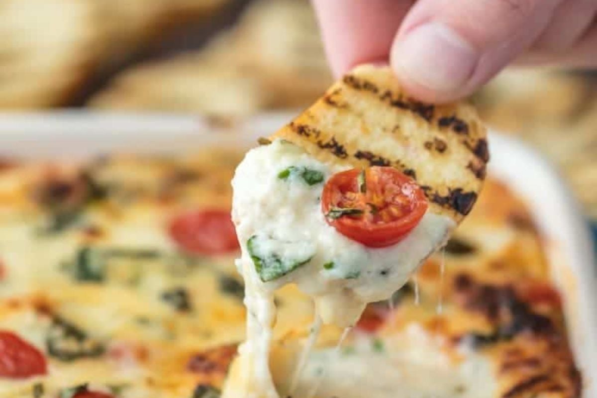 A hand holds a slice of flatbread, dipping it into a cheesy spinach and tomato dip from cheesy recipes, with melted cheese stretching between the dip and the bread.