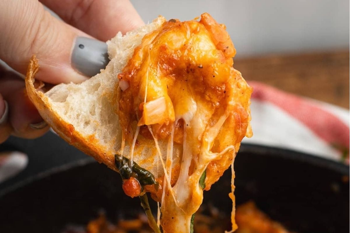 A hand pulling a slice of cheesy, tomato-based dish from a skillet, showing melted cheese stretching from the pan and highlighting one of many cheesy recipes.