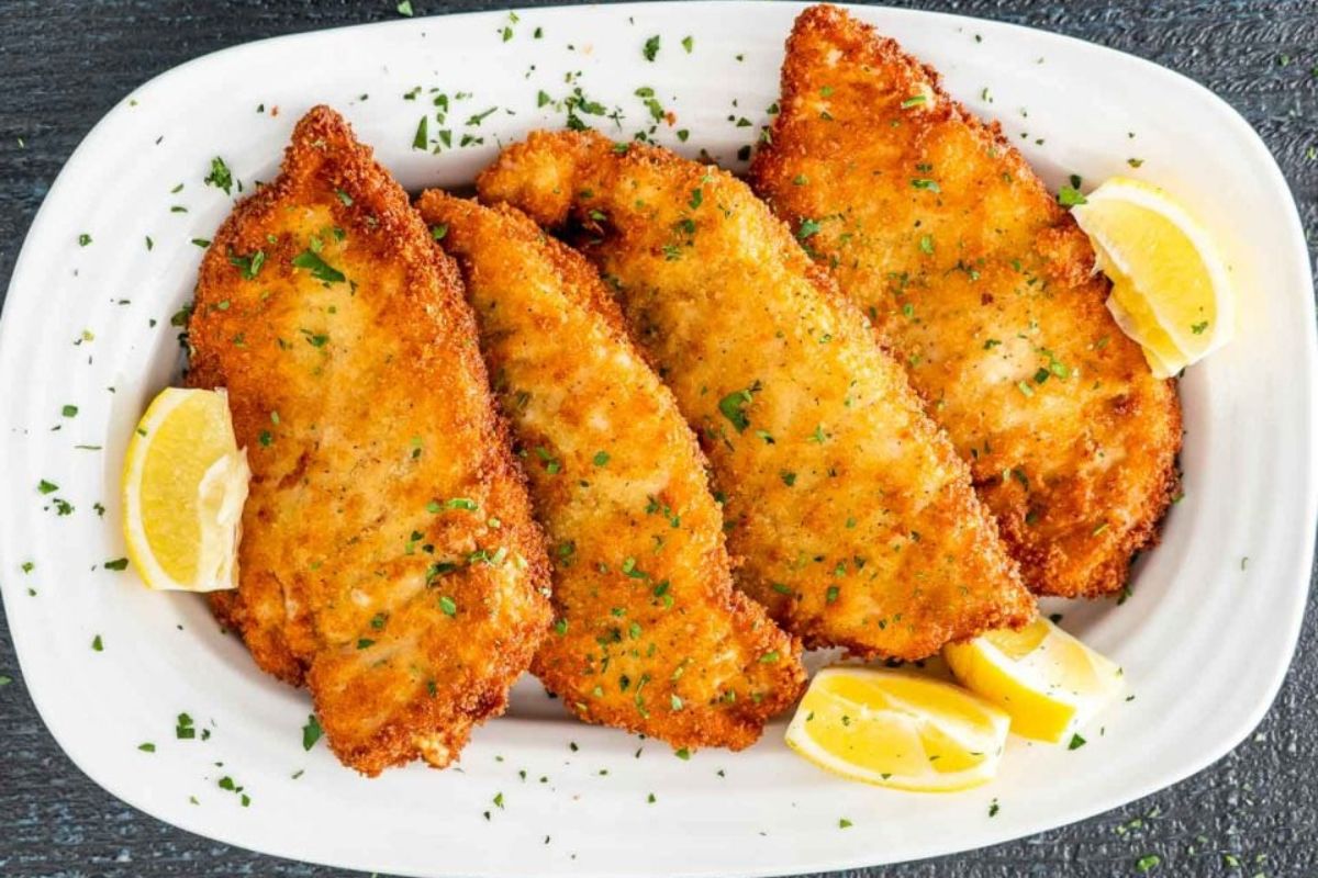 Plate with four breaded and fried fish fillets, garnished with chopped parsley and lemon wedges.