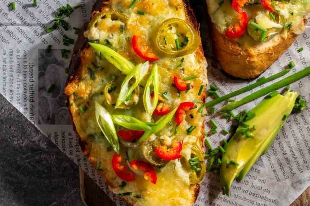 Two slices from our cheesy recipes toast topped with sliced avocado, green onions, and red chili, served on a newspaper-themed napkin.