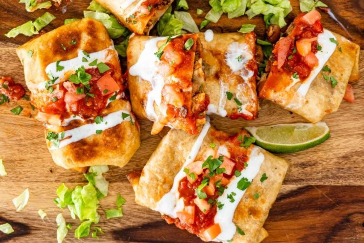 Four burrito recipes, grilled and topped with sour cream and salsa, garnished with cilantro, served on a wooden board with lime wedges and lettuce.